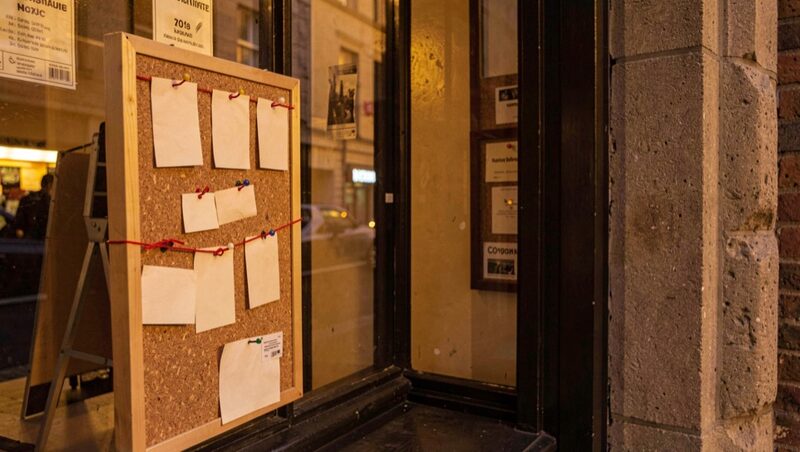 A cork noticeboard beside a theatre box office window holding blank pinned cards and red string, warm evening light